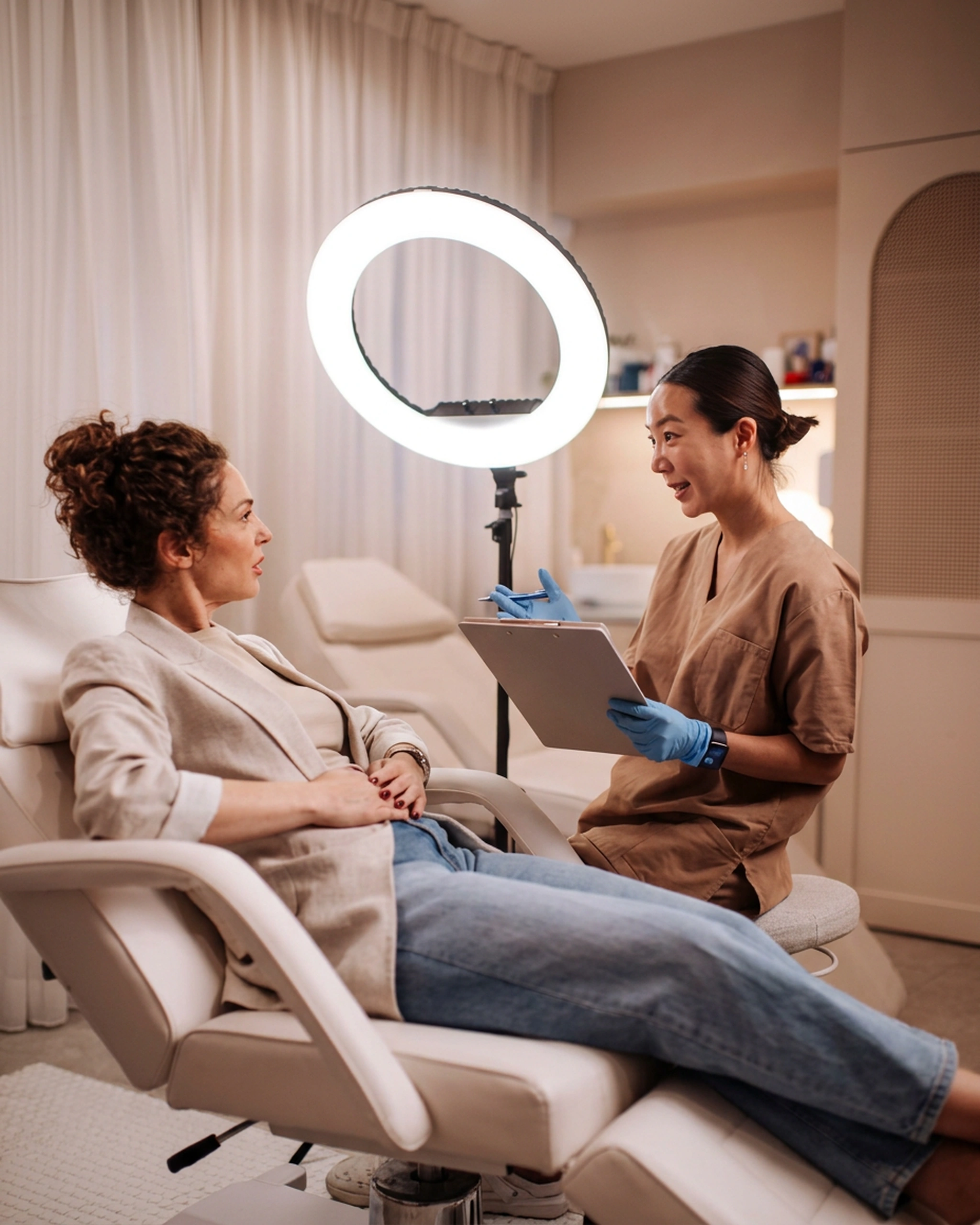 Med spa practitioner in scrubs reviewing a treatment plan on a clipboard with a client seated in a treatment chair in a bright aesthetic clinic